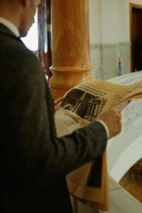 An elegant businessman in a suit reading a newspaper indoors near a marble column.