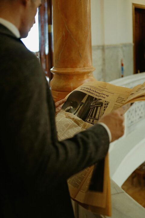 An elegant businessman in a suit reading a newspaper indoors near a marble column.